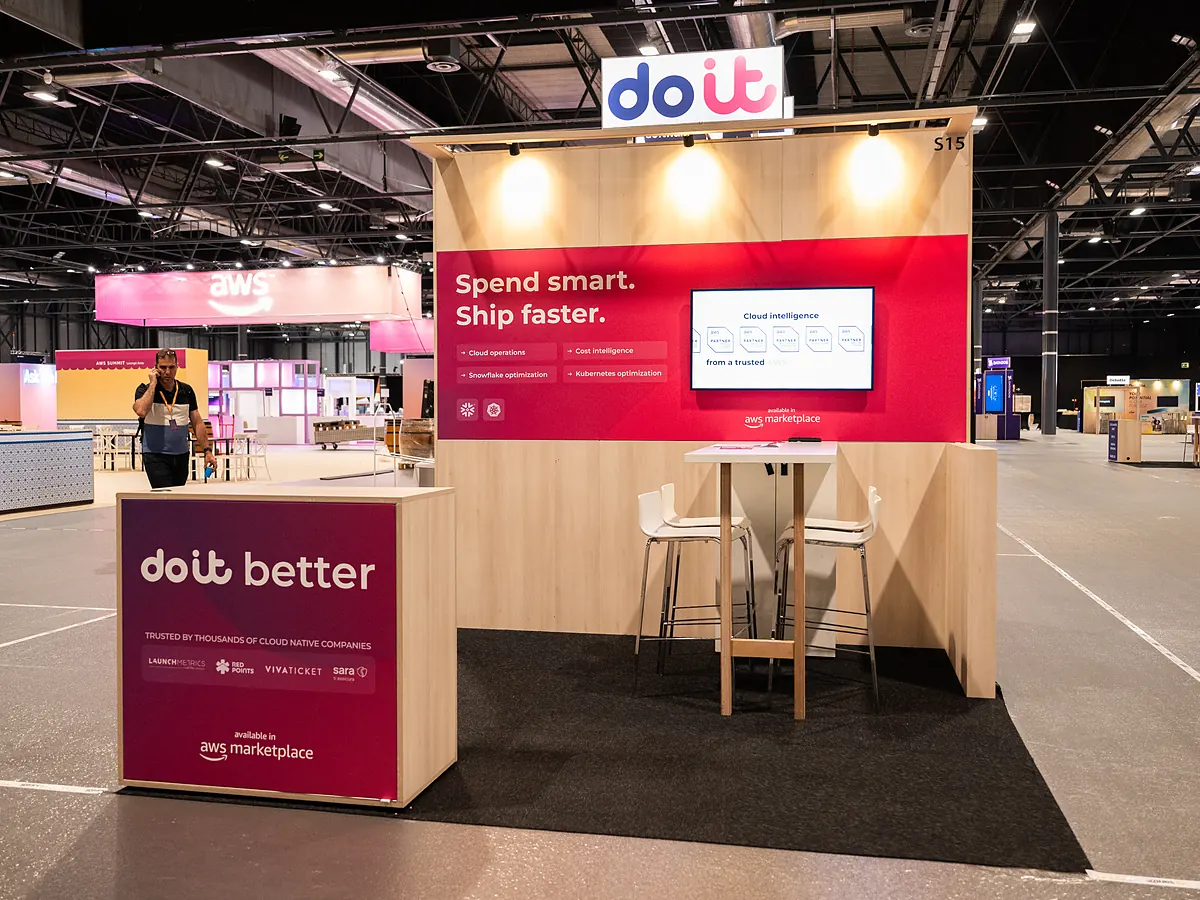 Stand van DoiT met een rode printwand, een scherm, een statafel met krukken en een balie met logo