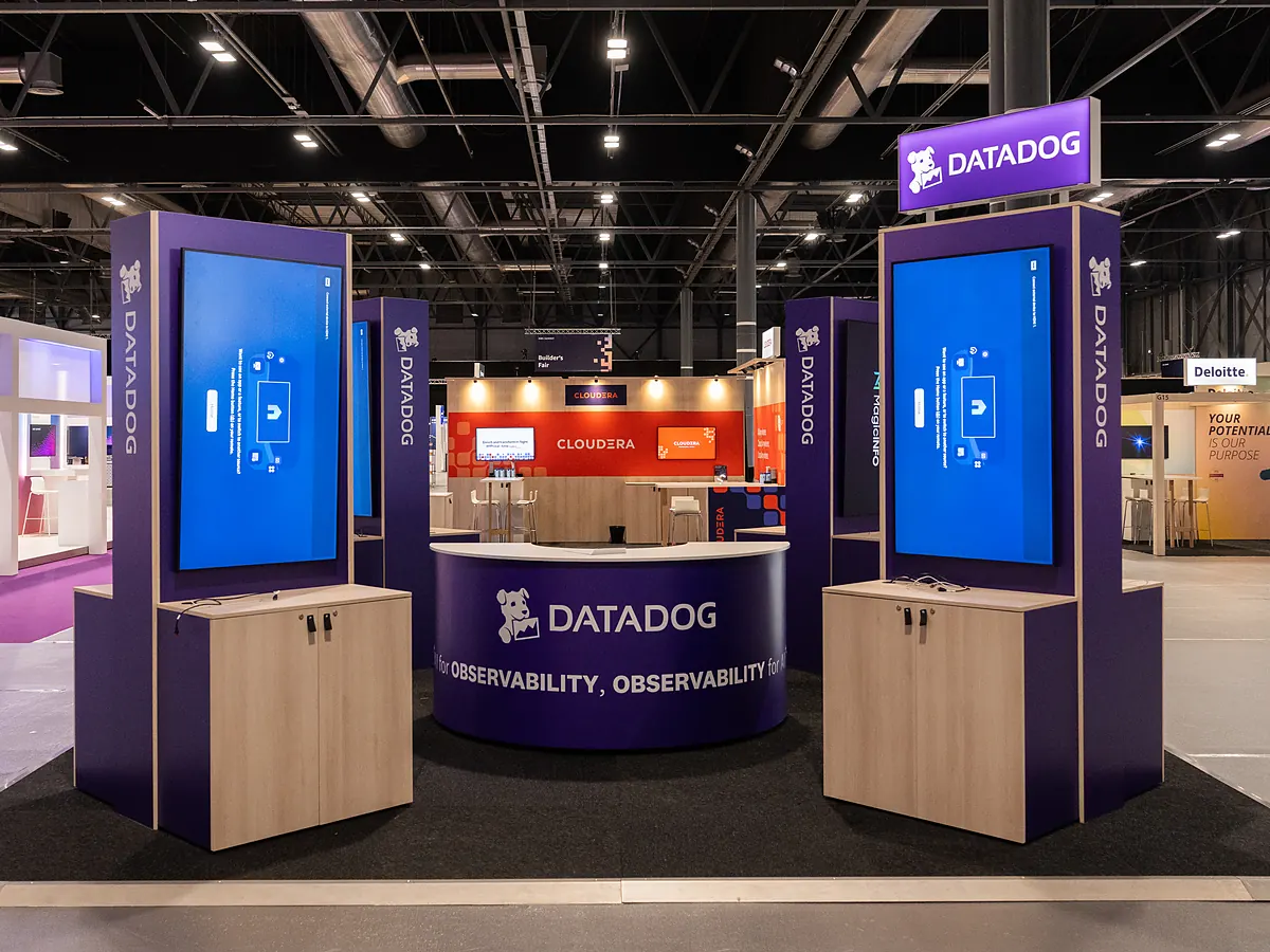 Stand van Datadog met twee paarse torens met schermen en een ronde balie met het logo